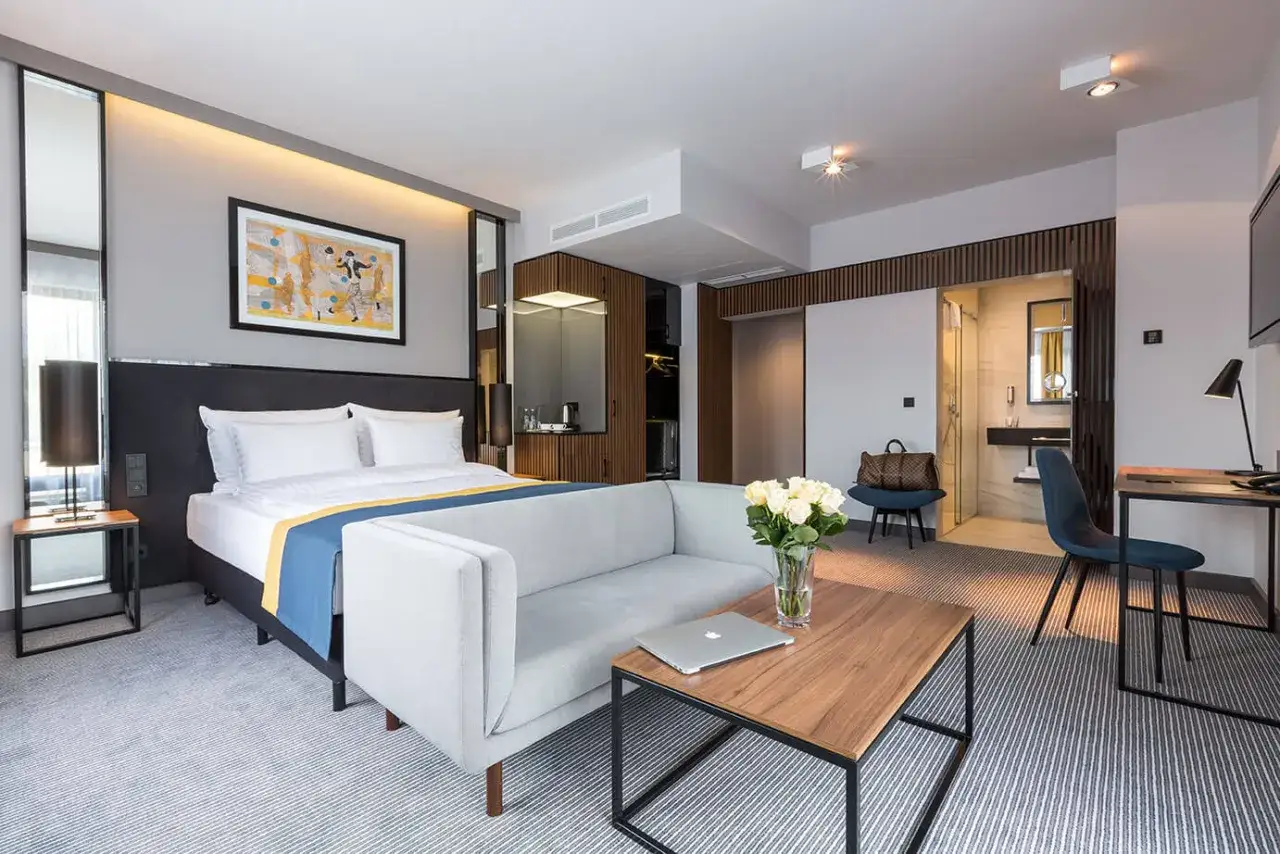 Elegancki pokój hotelowy z wygodnym łóżkiem, sofą i stolikiem kawowym. W tle widać łazienkę i aneks kuchenny. Idealne miejsce na odpoczynek po podróży, jak w hotelu Ibis.