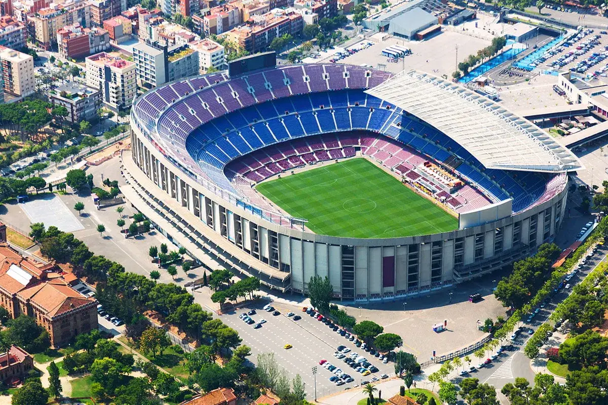 Camp Nou zwiedzanie: imponujący stadion piłkarski z zieloną murawą i niebiesko-bordowymi trybunami, otoczony miejską zabudową i drzewami.
