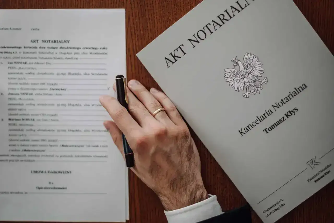 Akt notarialny dotyczący darowizny mieszkania. Dłoń z obrączką trzyma długopis nad dokumentem.