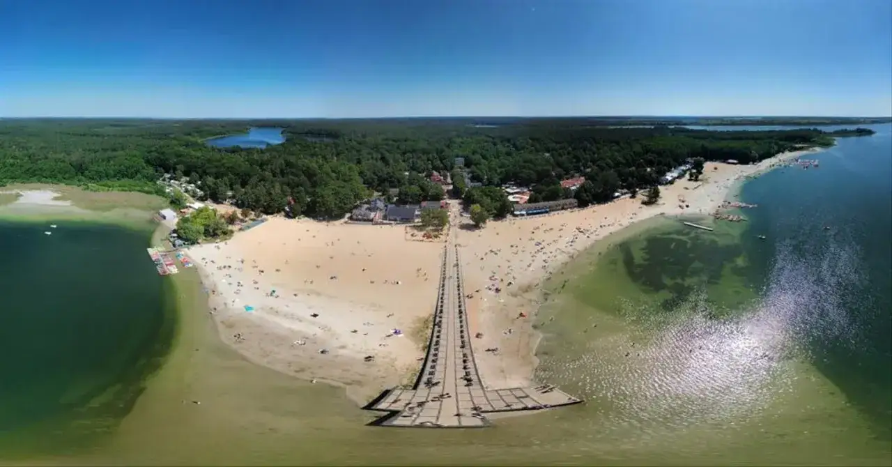 Skorzęcin atrakcje: plaża pełna ludzi, molo, las i jezioro w słoneczny dzień.