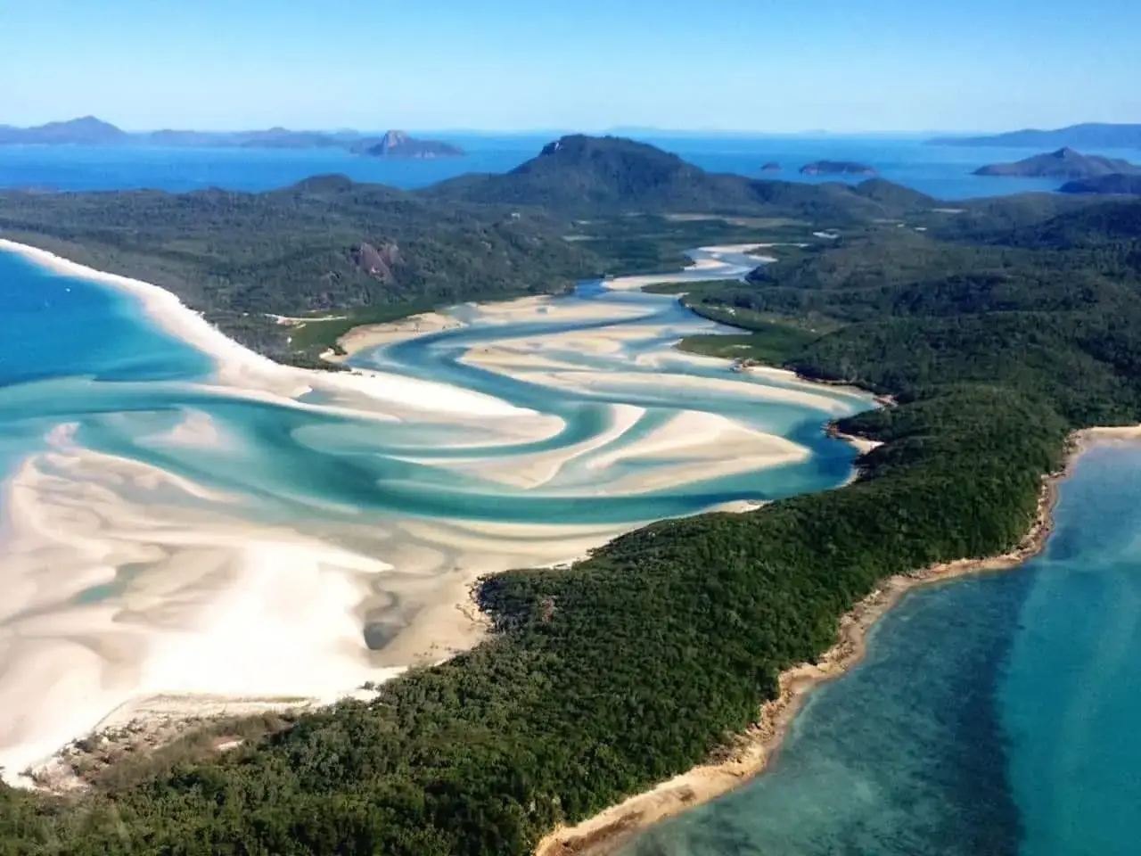 Widok z lotu ptaka na Whitehaven Beach z turkusową wodą i białym piaskiem tworzącymi wiry. W tle zielone wzgórza i wyspy.