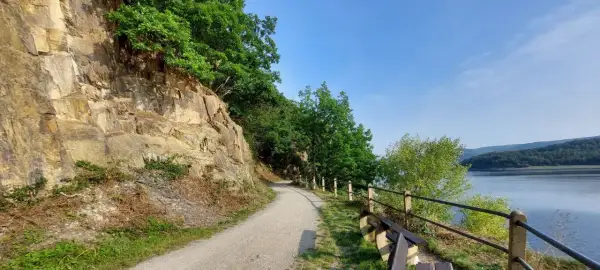 Ścieżka wzdłuż skalnego zbocza, z widokiem na jezioro i las. Krajobraz przypomina Eifel.