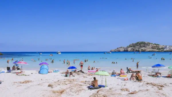 Słoneczna plaża Cala Agulla tętni życiem. Ludzie kąpią się w krystalicznie czystej wodzie, opalają na piasku i odpoczywają pod parasolami.