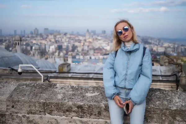 Kobieta w jasnoniebieskiej kurtce i okularach przeciwsłonecznych na tle panoramy Stambułu.