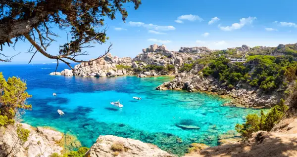 Turkusowa woda zatoki na Costa Smeralda, łodzie kołyszące się na falach, skaliste wybrzeże porośnięte zielenią.
