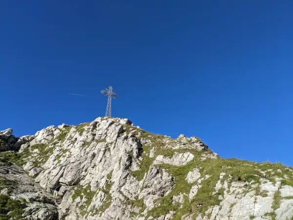 Na szczycie skalistego zbocza, porośniętego trawą, stoi metalowy krzyż. W tle błękitne niebo, a w oddali widać samolot. To widok z Giewontu, który przypomina, ile ma wysokości.