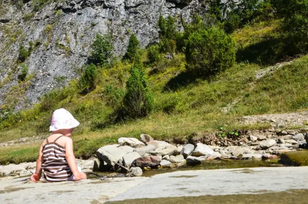Dziecko w kapelusiku siedzi nad strumieniem w Małych Pieninach, otoczone zielenią i skalistym zboczem.