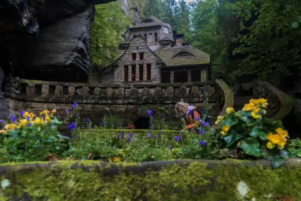 Kamienny domek w otoczeniu skał i zieleni, przypominający baśniową scenerię czeskiej Saksonii. Kobieta pochyla się nad kwiatami.