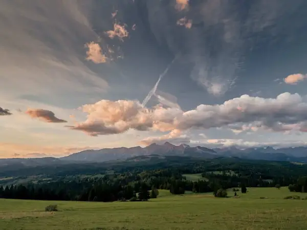 Widok na Tatry z zielonej łąki, zalesione wzgórza i chmury na niebie.