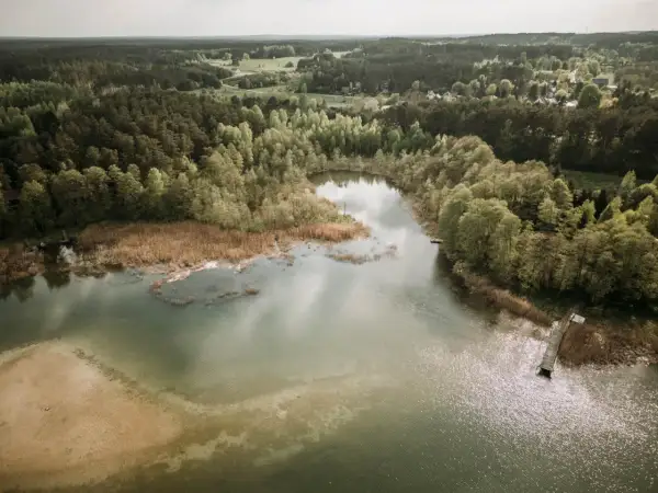 Widok z lotu ptaka na spokojne jezioro otoczone lasem, z piaszczystym brzegiem i drewnianym pomostem.
