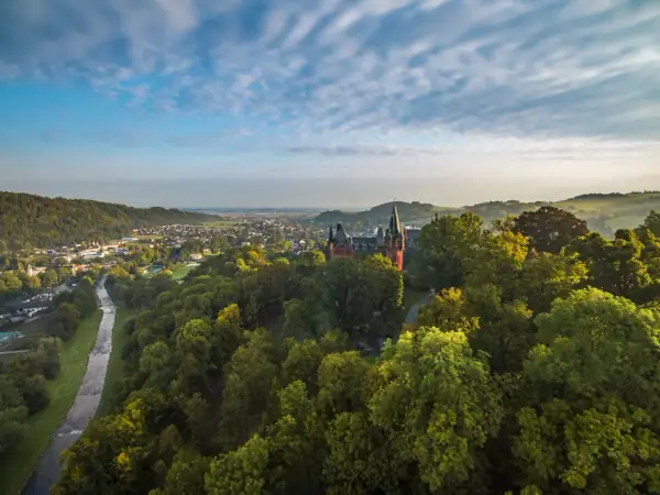Widok na zamek w Hradcu nad Moravicí otoczony zielonymi drzewami, z miastem i rzeką w tle.