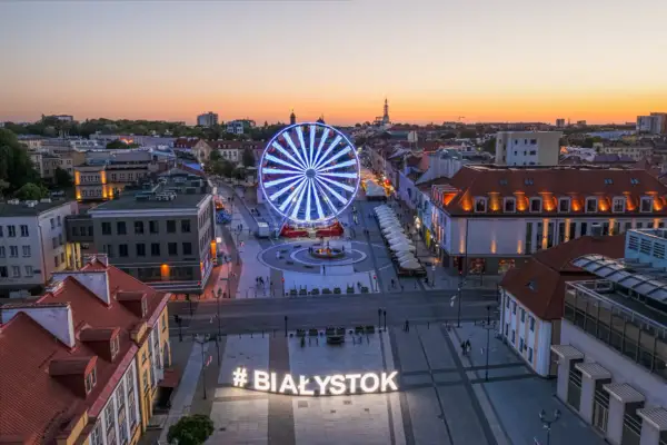 Widok z góry na plac z podświetlanym napisem #BIAŁYSTOK i diabelskim młynem o zachodzie słońca.